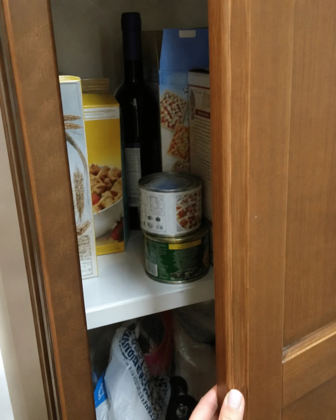Kitchen pantry cabinet slightly open with a wine bottle tucked behind cereal boxes, a hand pushing it back