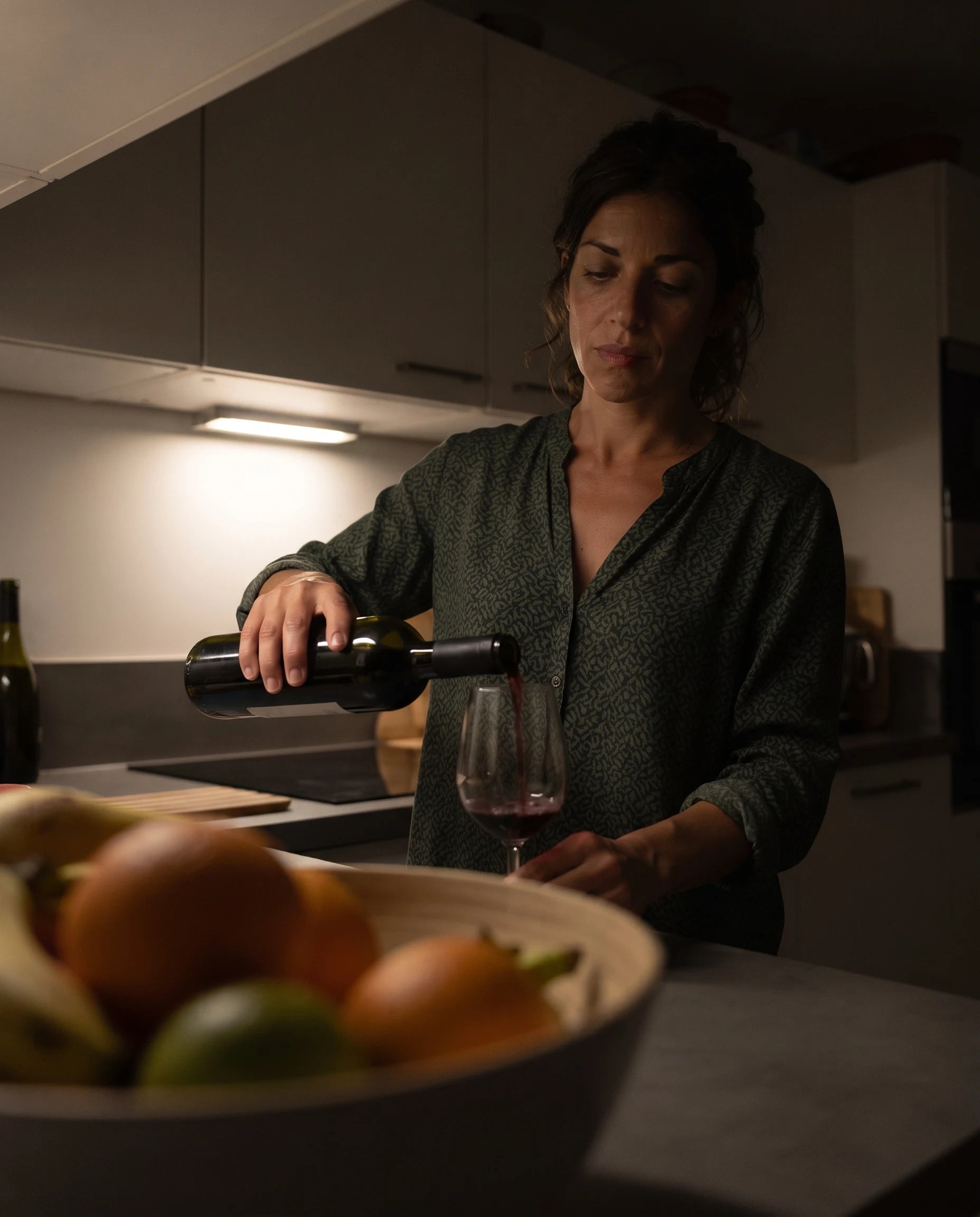 Woman pouring wine alone in a dim kitchen late at night, phone propped on the counter filming candidly