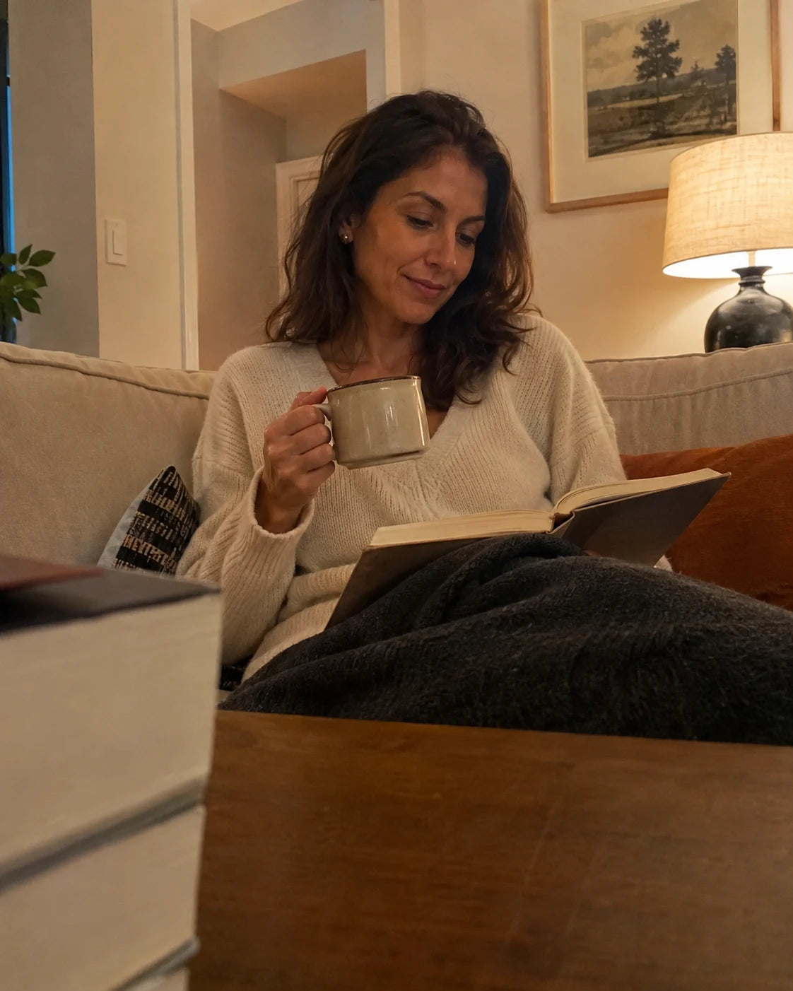 Woman relaxed on her couch in the evening with tea and a book, no wine, candid phone-propped shot