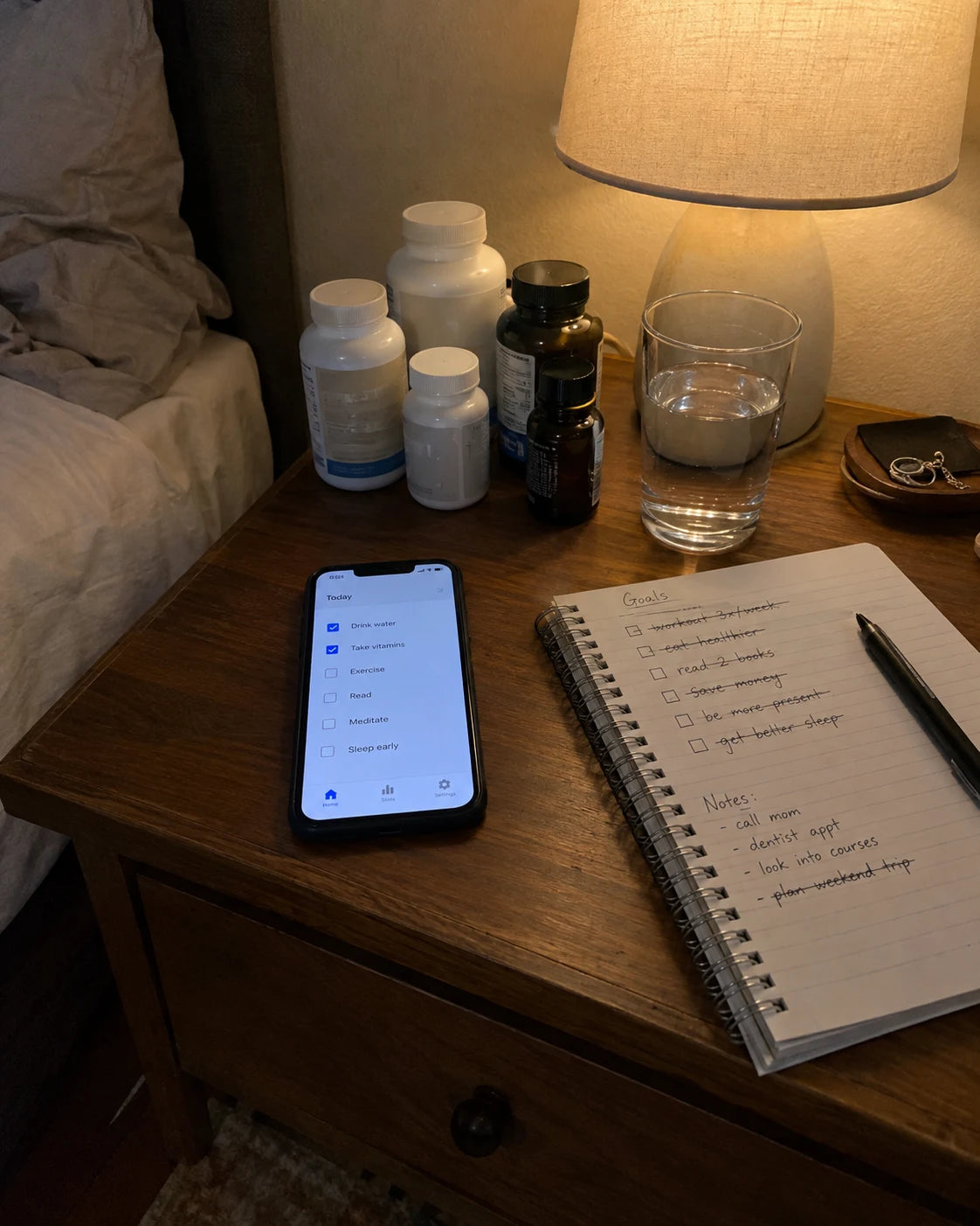 Nightstand with supplement bottles, a phone showing a habit-tracking app, and a notebook of crossed-out notes
