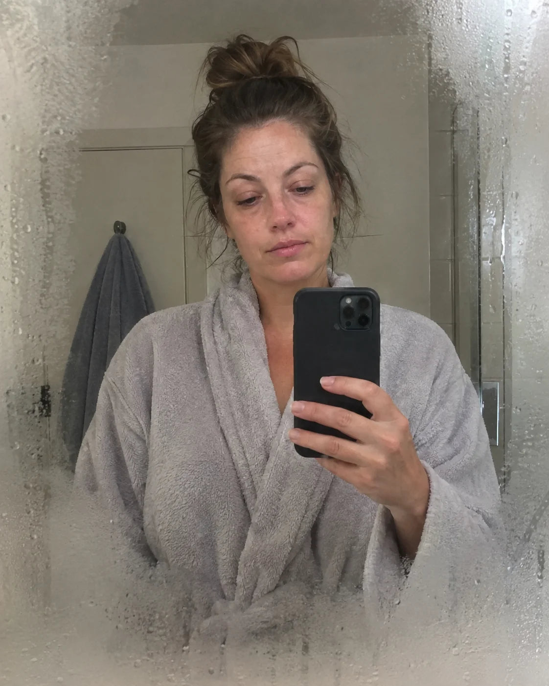A woman's tired, puffy morning face reflected in a steamed-up bathroom mirror, taking a selfie