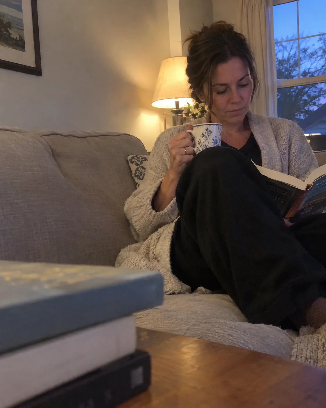 A woman relaxed and content on her couch in the evening, reading a book and holding a warm cup of tea