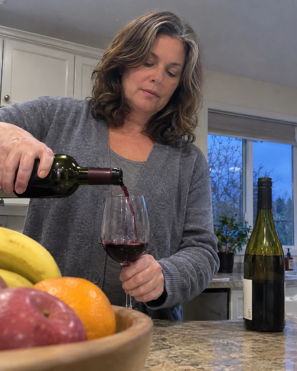 A woman in a kitchen at dusk pouring wine into a glass, a half-empty bottle beside her, filmed from a phone propped against a fruit bowl on the counter