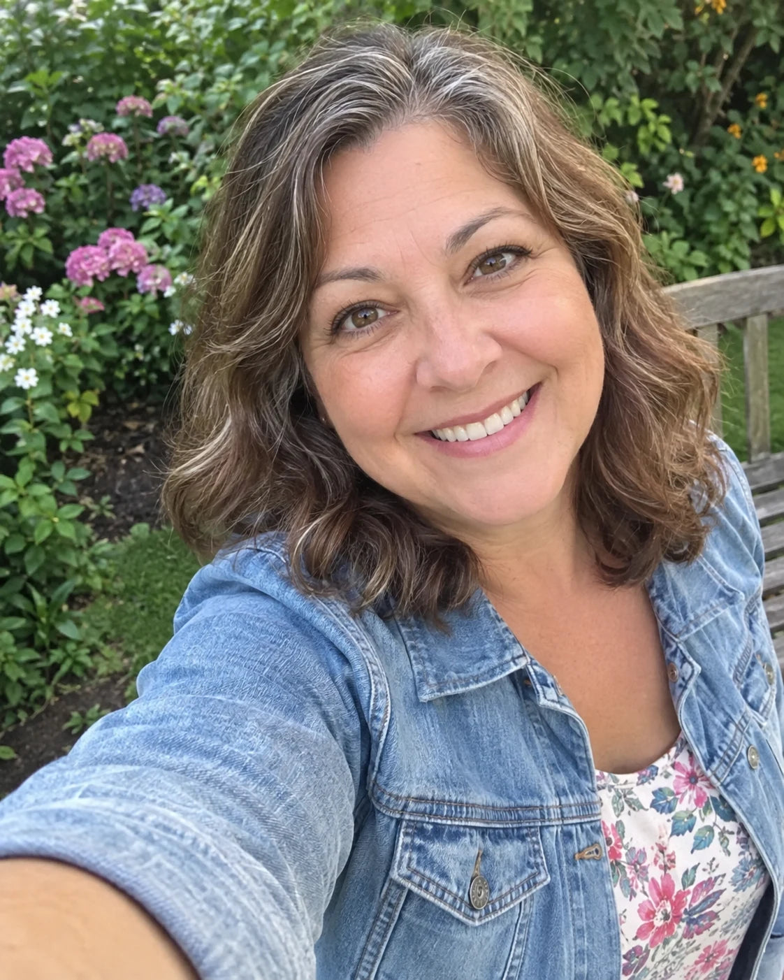 A woman taking a selfie outdoors in a garden, radiant genuine smile, arm extended toward the camera, flowers in the background
