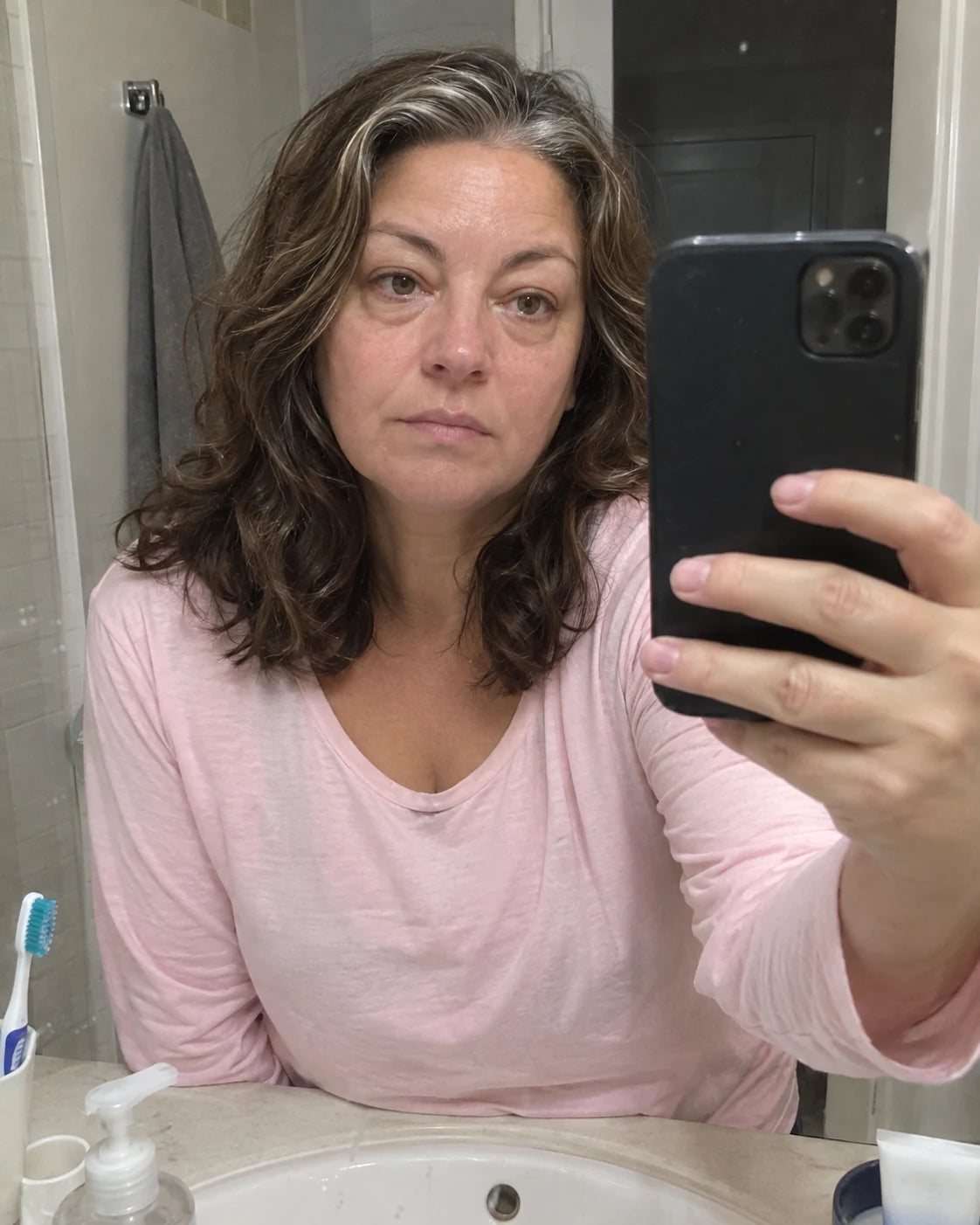 A woman taking a tired morning mirror selfie in a bathroom, phone visibly held in her extended hand