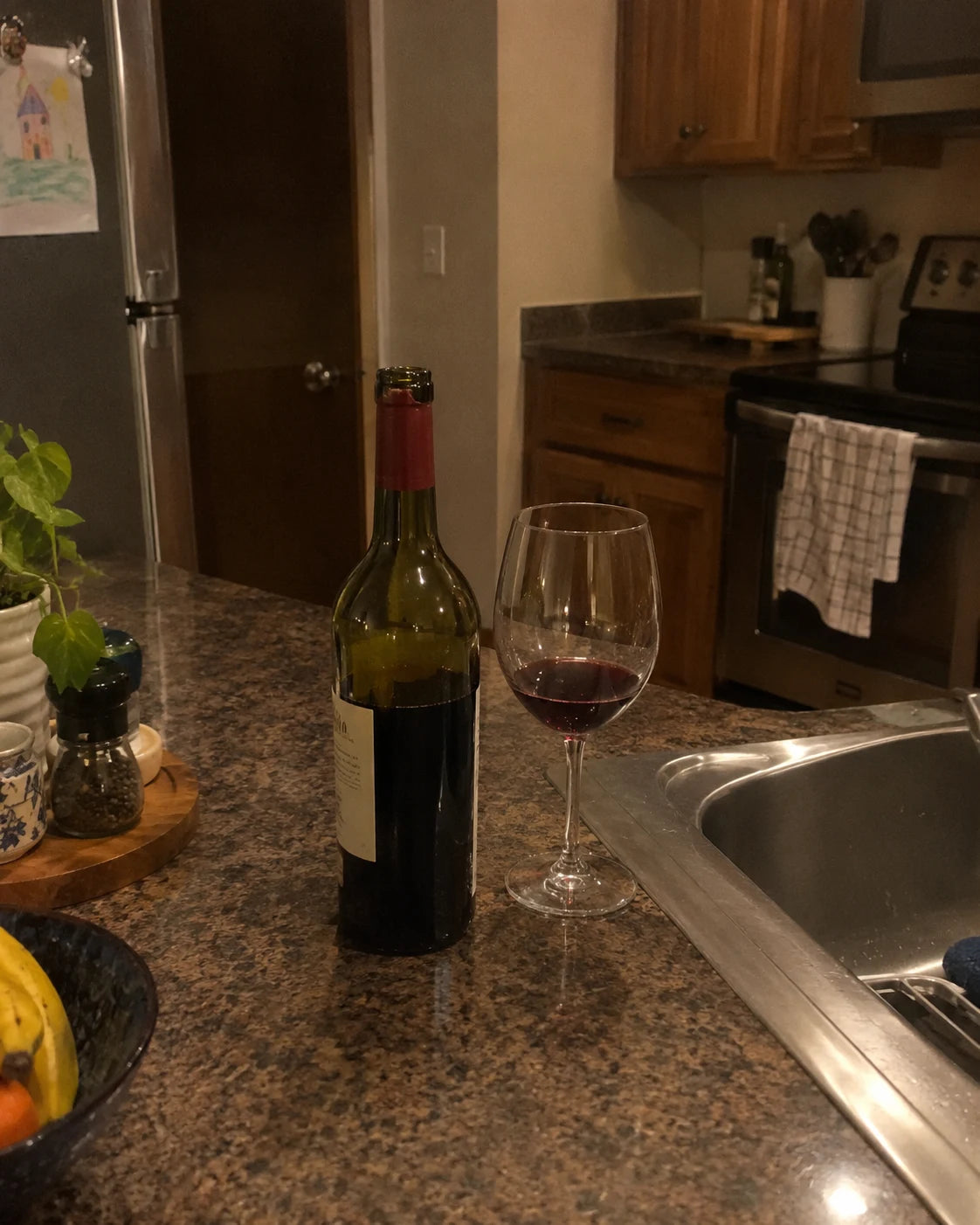 A half-empty wine bottle and glass on a kitchen counter at night
