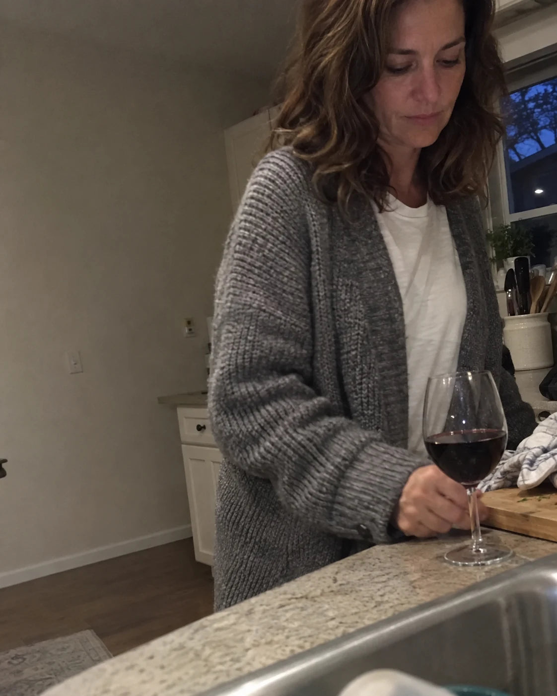 A glass of red wine already poured on a kitchen counter at dusk, phone propped nearby