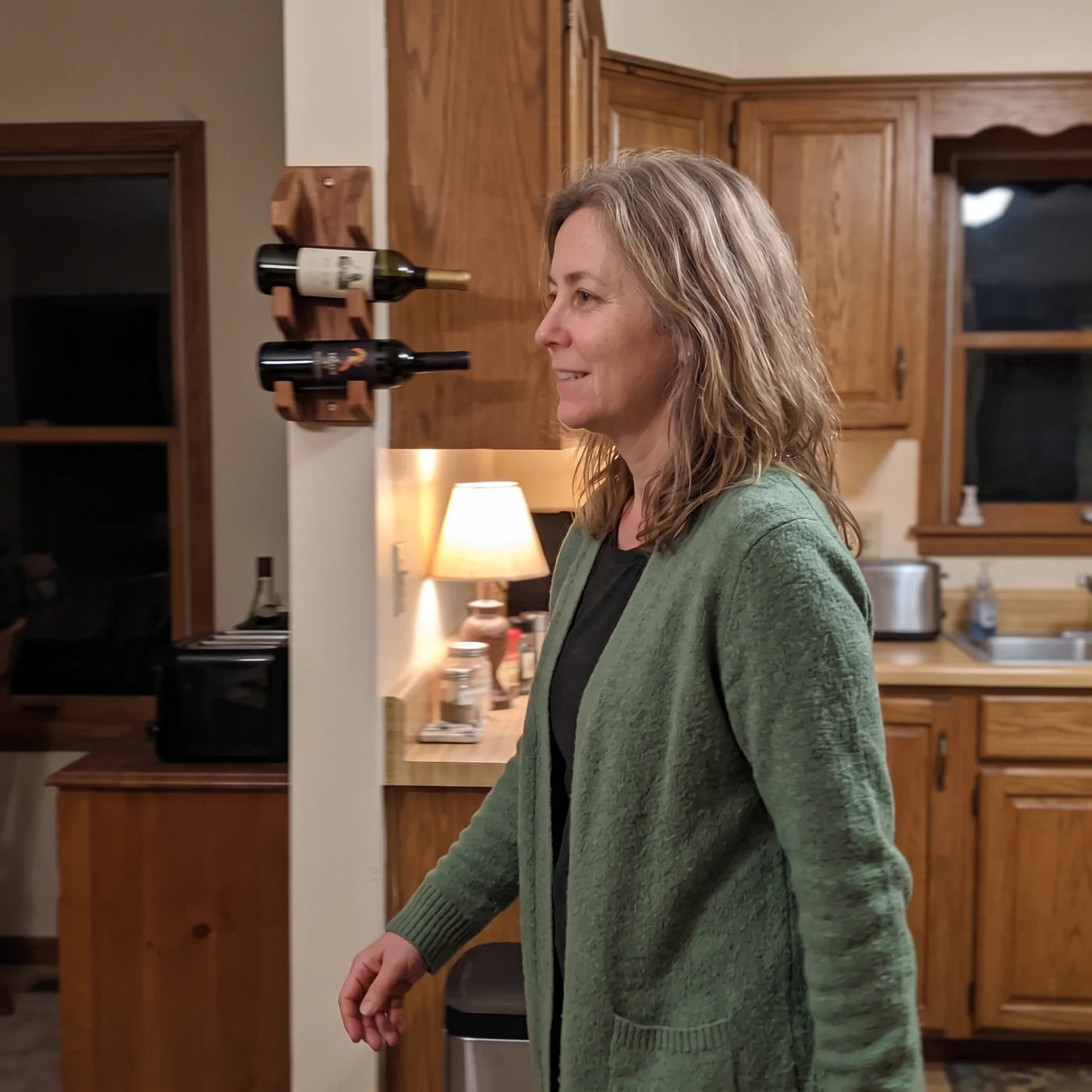 Woman walking calmly through her kitchen in the evening, passing a wall-mounted wine rack without reaching for it