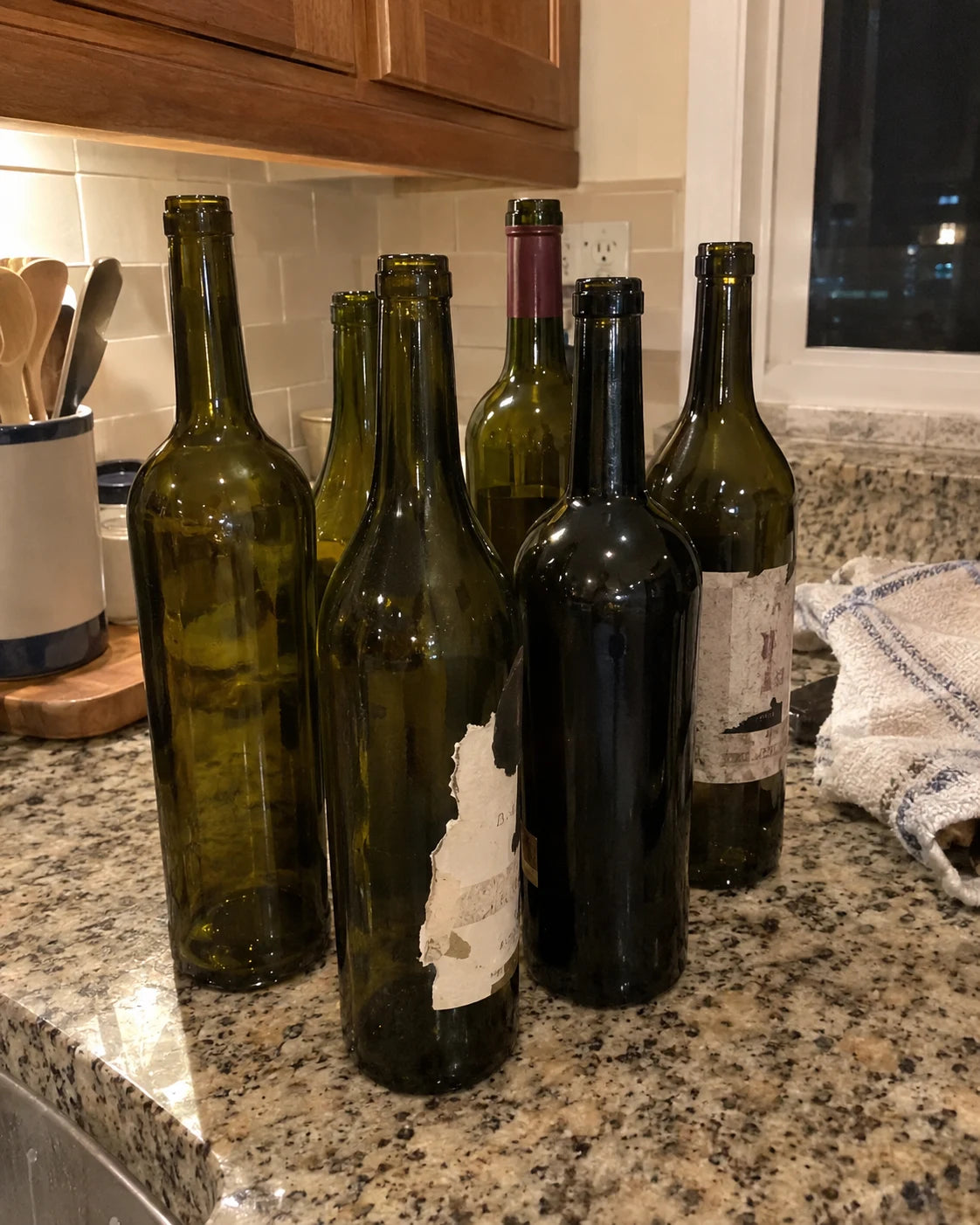 Several empty wine bottles with blank labels lined up on a kitchen counter at night