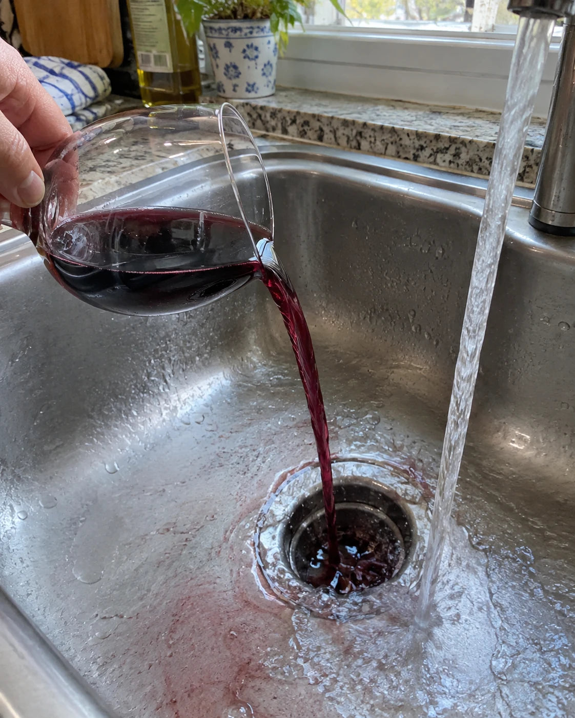 Red wine being poured out of a glass into a kitchen sink drain
