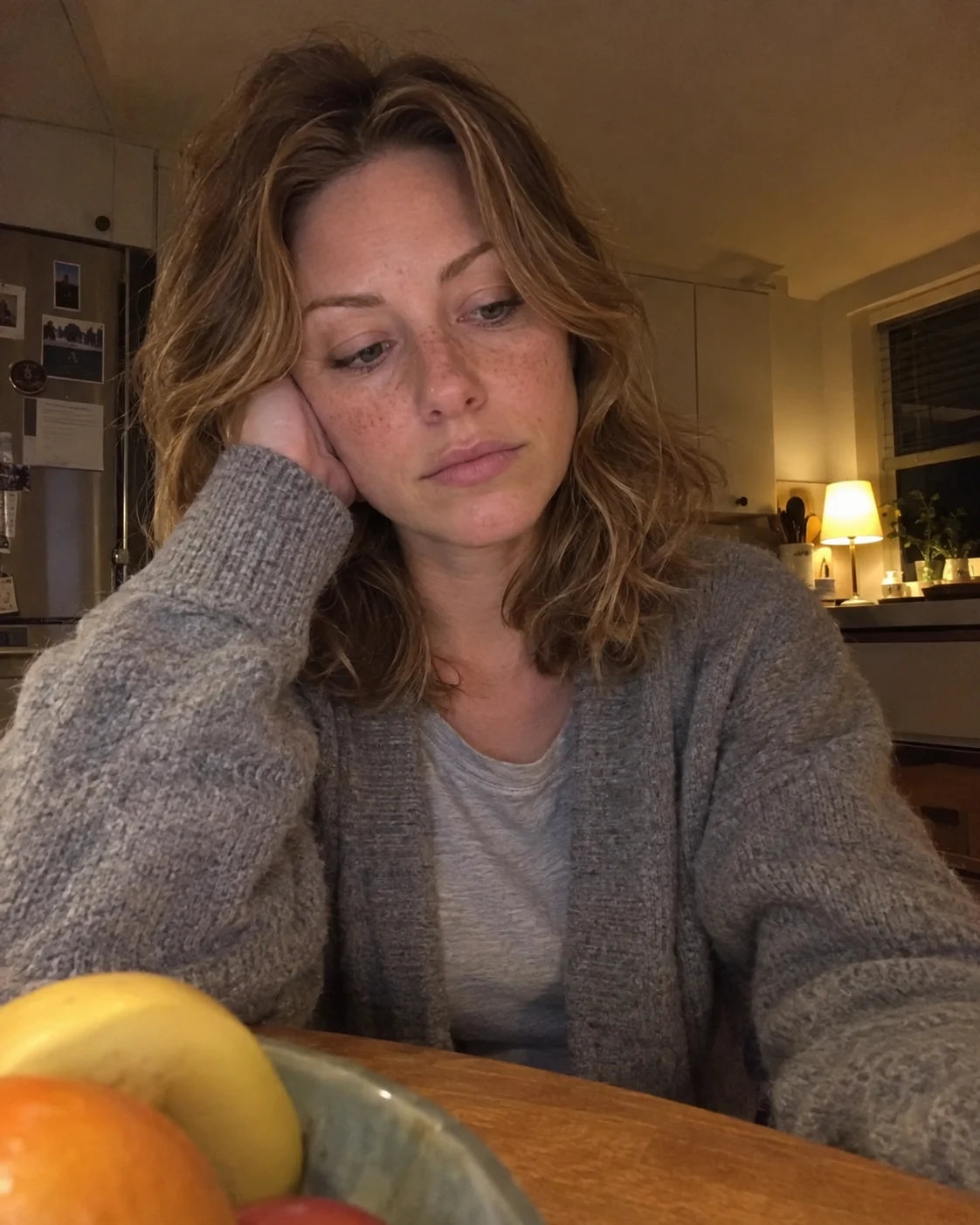 A woman sitting alone at a kitchen table late at night, tired, resting her head on her hand