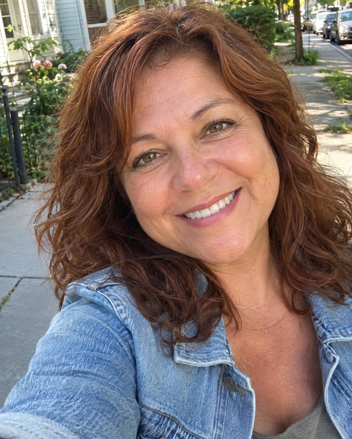 A close selfie-style portrait of a woman smiling radiantly outdoors in the morning sunlight