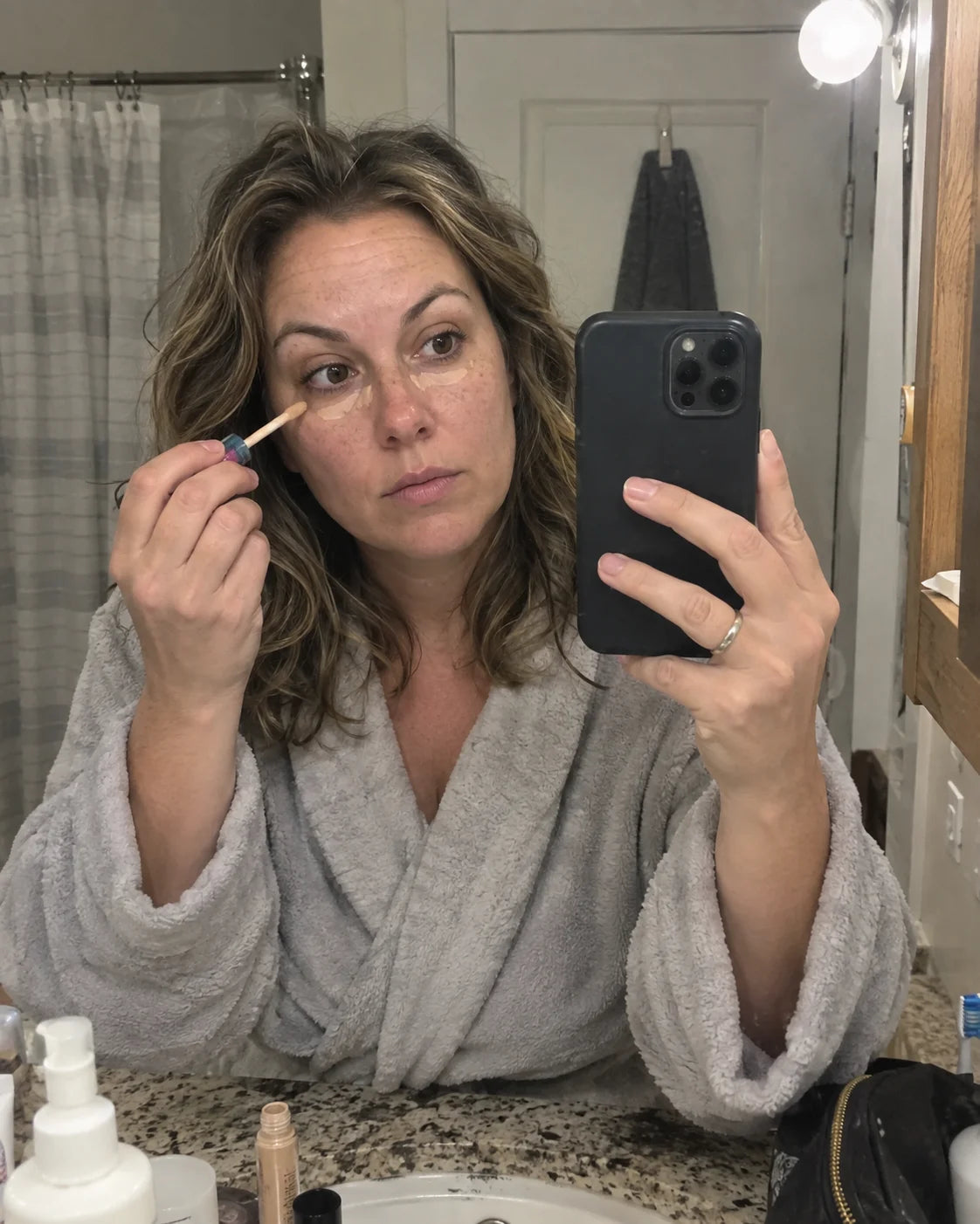 Woman applying concealer under her eyes at the bathroom mirror in the morning