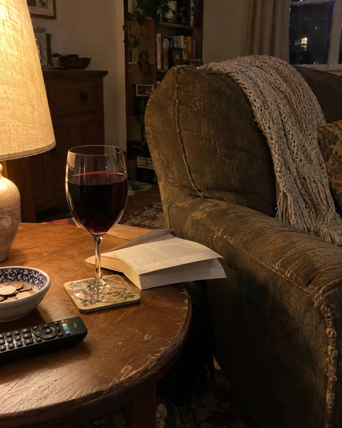A glass of wine on a side table beside a lamp and an open book in a quiet living room at night