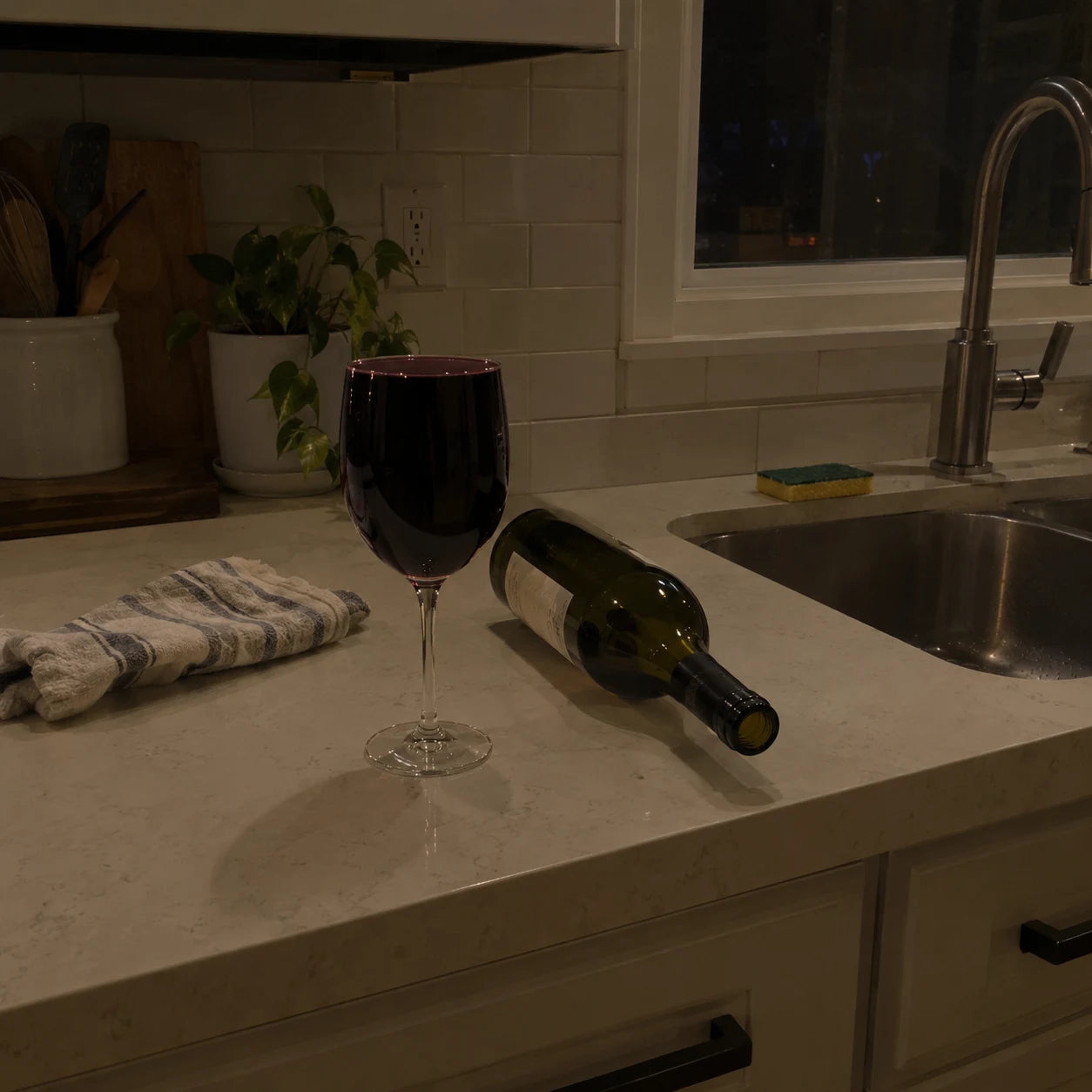 A full glass of wine next to an empty bottle set down on a kitchen counter at night
