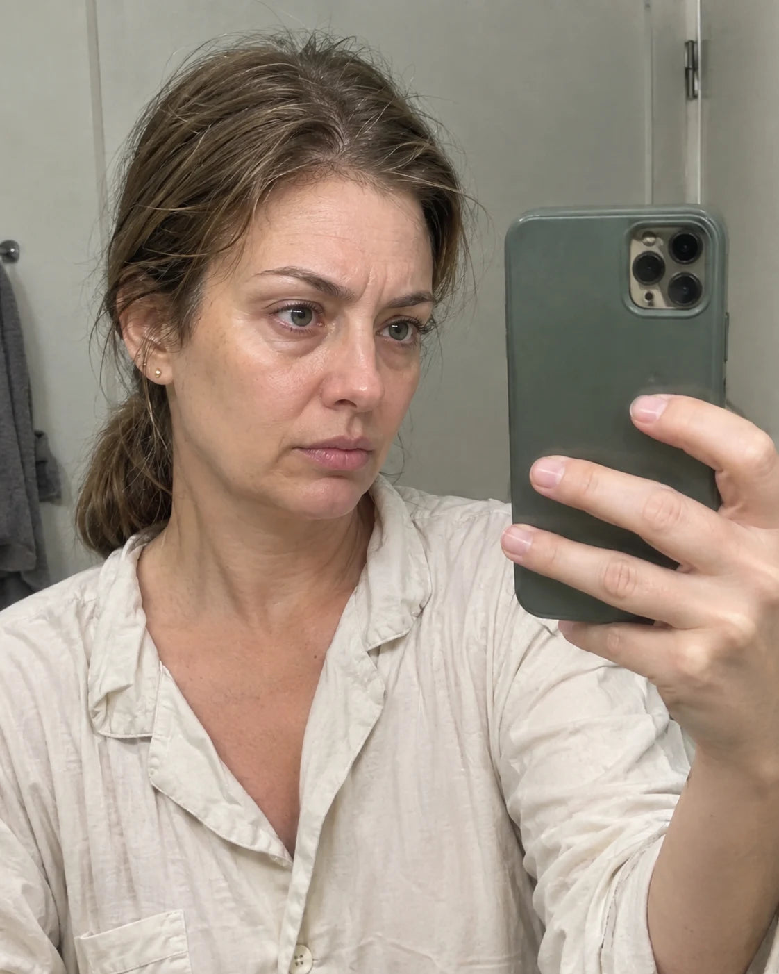 A woman taking a tired mirror selfie, examining her own puffy face closely