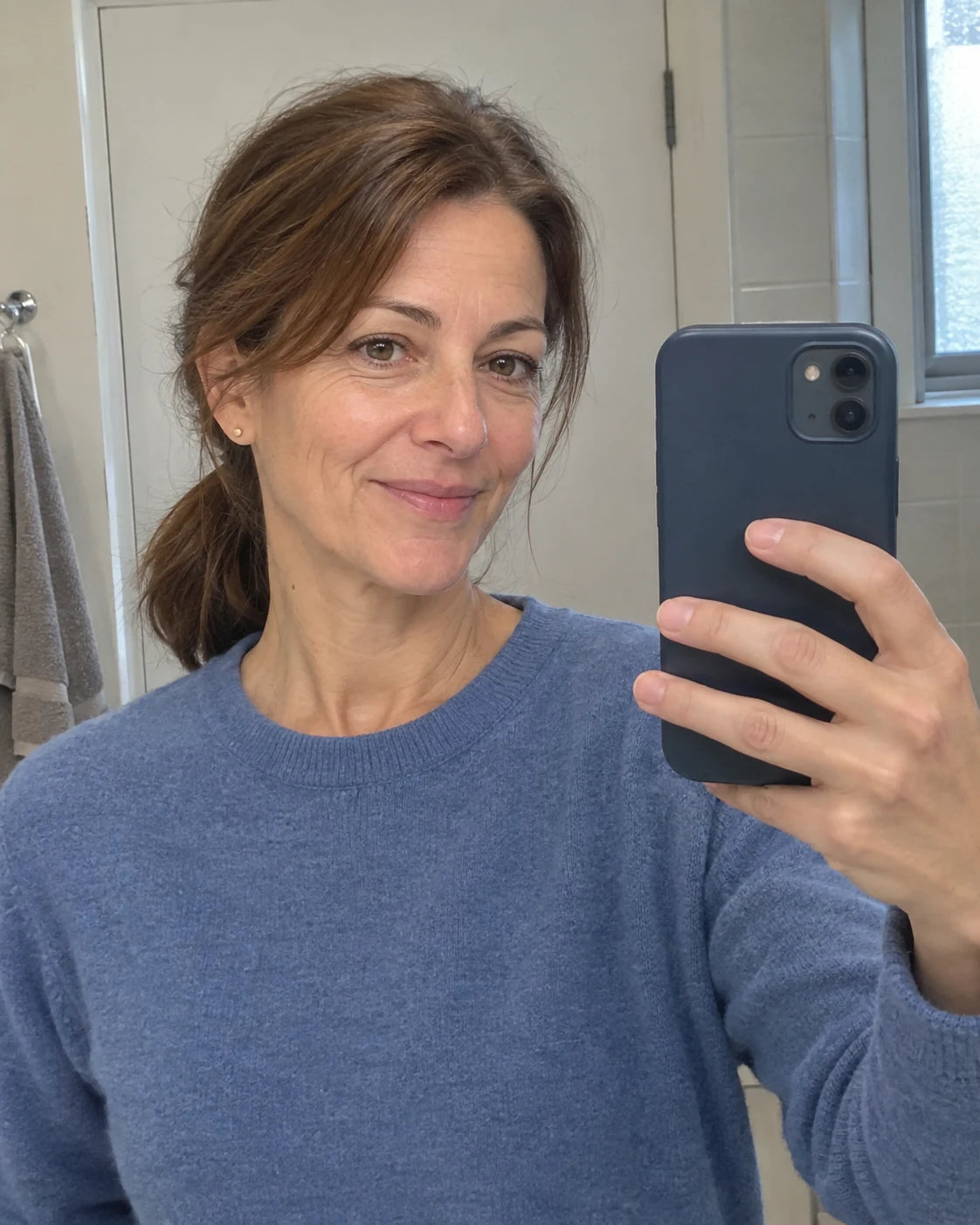The same woman taking a mirror selfie, relaxed and smiling, wearing a different sweater