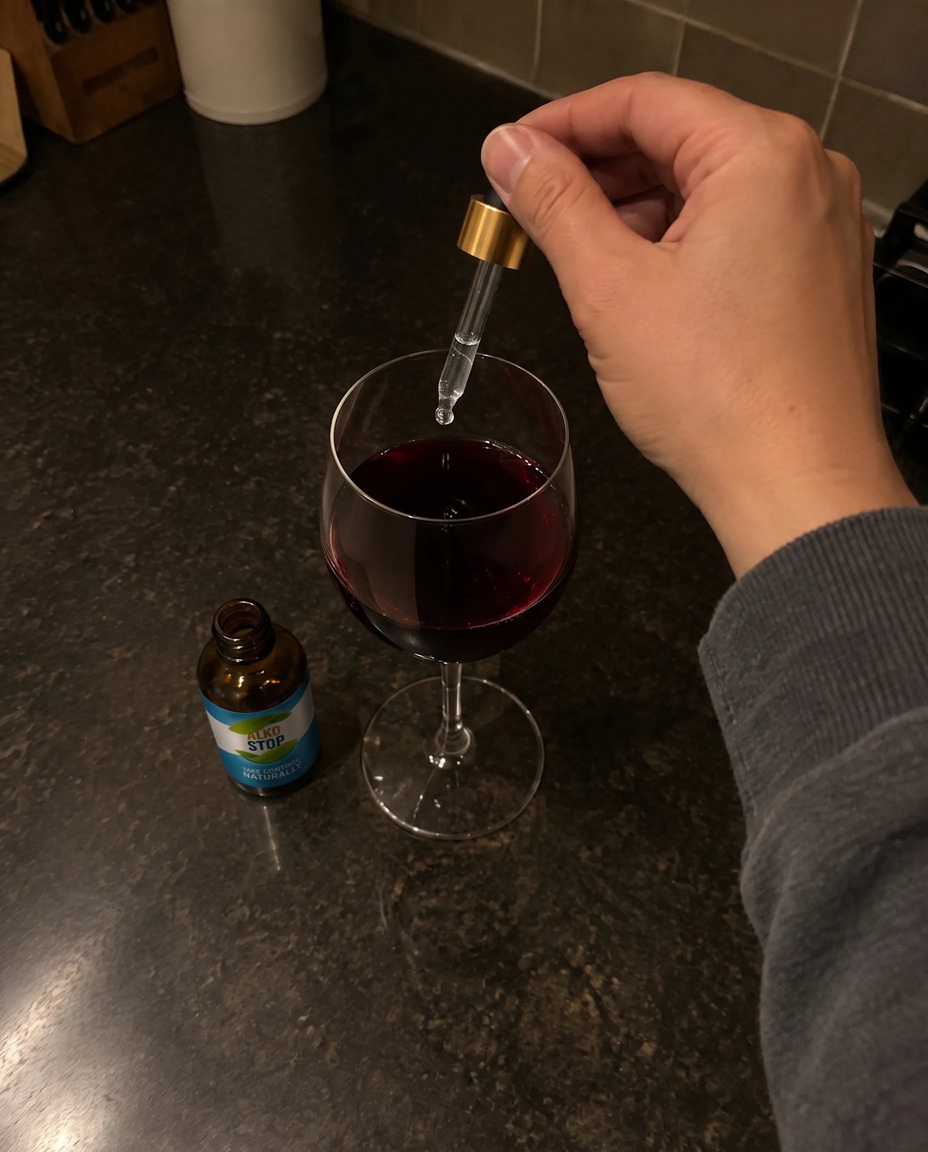 A hand adding a drop of AlkoStop into a glass of wine, with the open bottle sitting on the counter