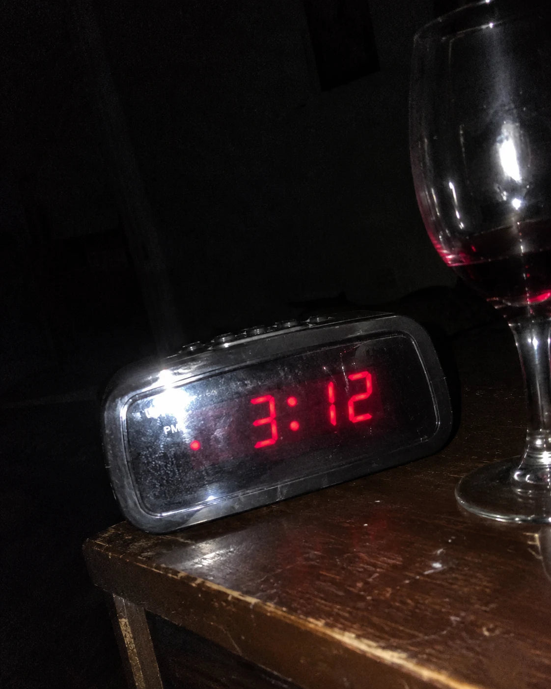 A digital alarm clock glowing 3:12 on a dark nightstand beside a half-full wine glass in a dim bedroom