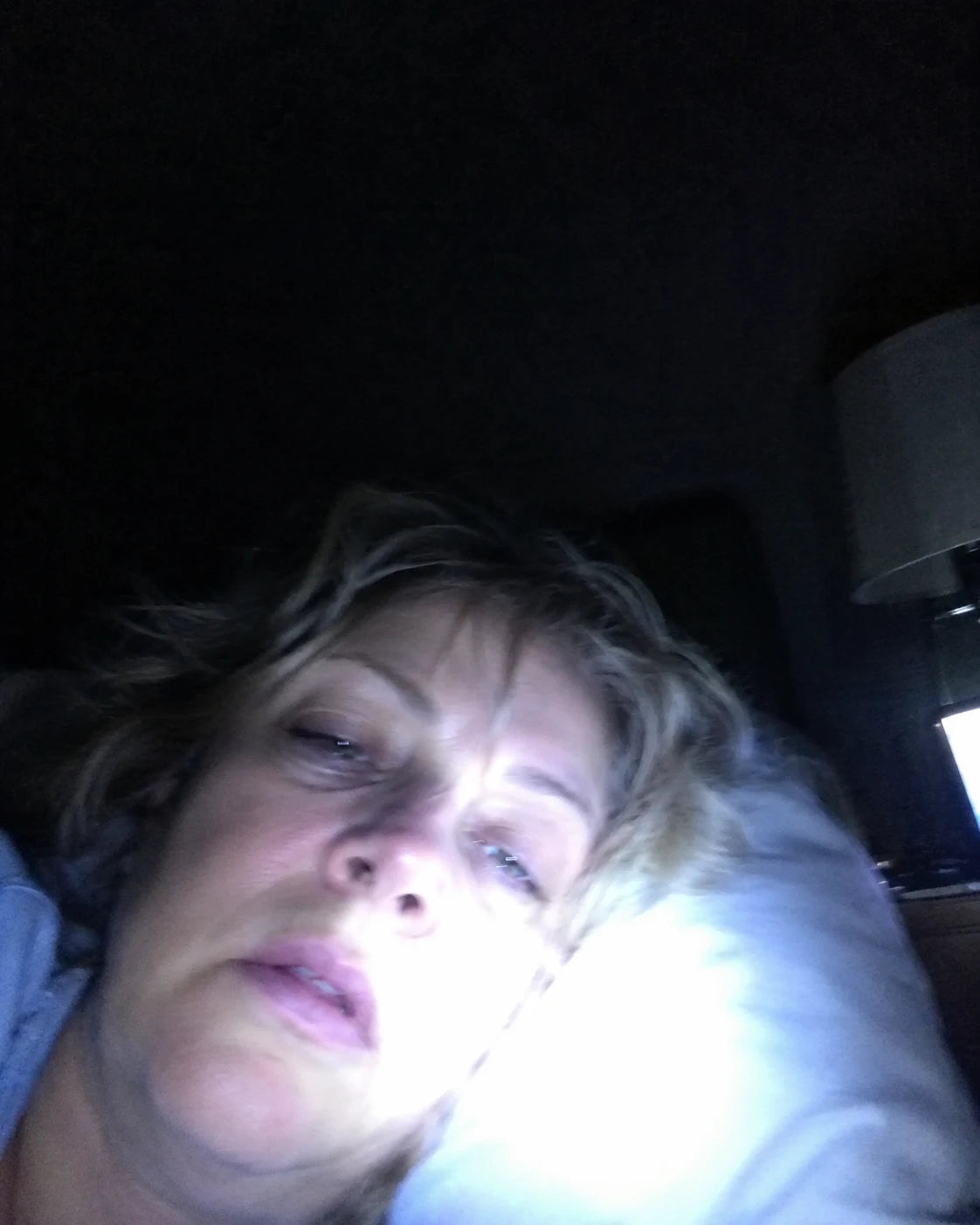 A tired woman lying awake in bed at night staring at the ceiling, lit only by a phone propped on the nightstand