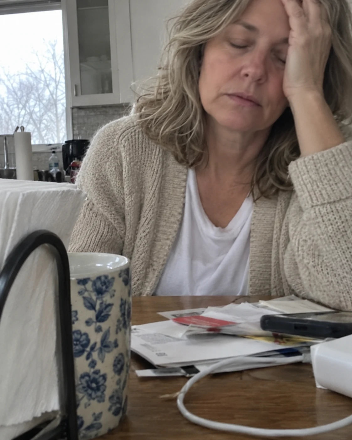 An exhausted woman resting her head on her hand at a kitchen table in the morning beside an untouched cup of coffee