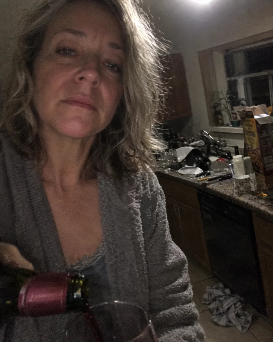 A tired woman taking a selfie while pouring a glass of red wine in a dim kitchen late at night