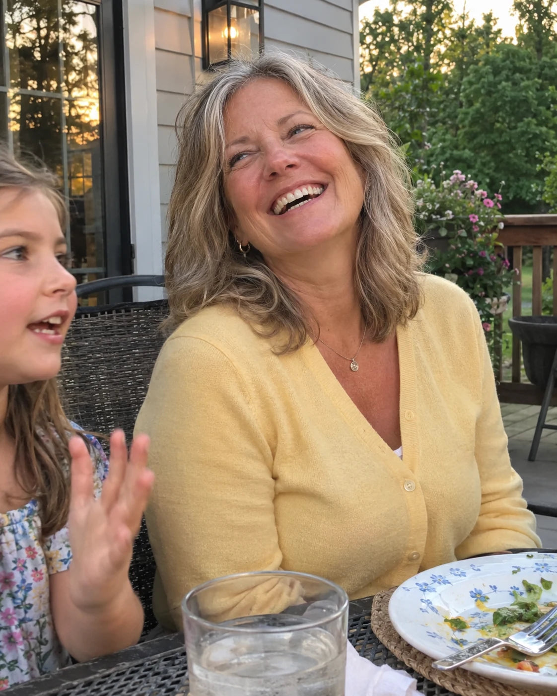 A smiling woman laughing at an outdoor family dinner table beside her young granddaughter