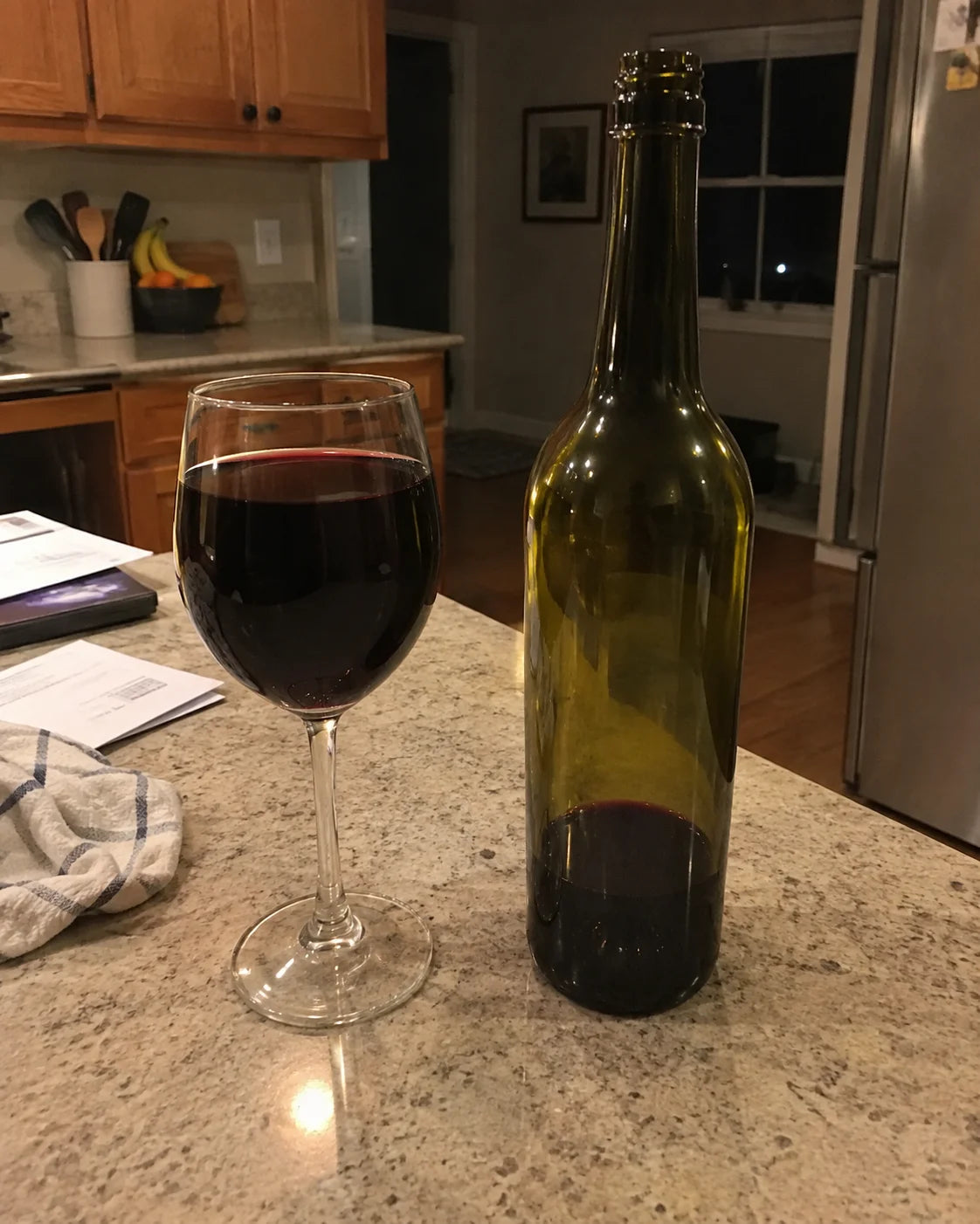 A full wine glass and a wine bottle, noticeably more empty than expected, on a kitchen counter in the evening