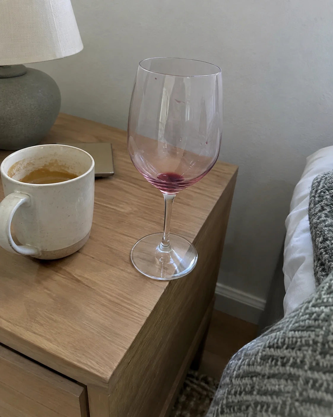 An empty wine glass on a nightstand next to a half-finished coffee mug in soft morning light
