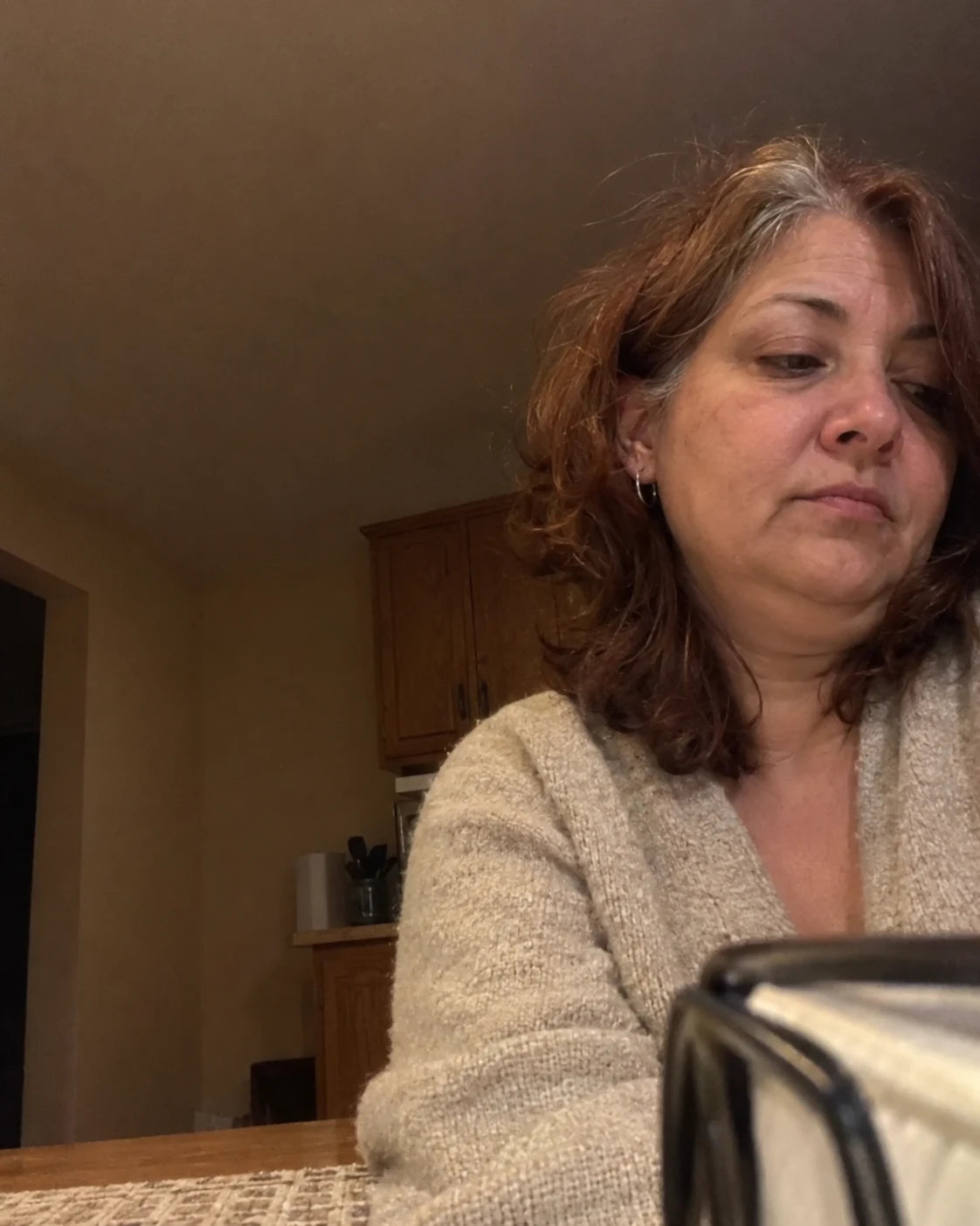 A tired woman sitting alone at her kitchen table in the evening, phone propped against a napkin holder