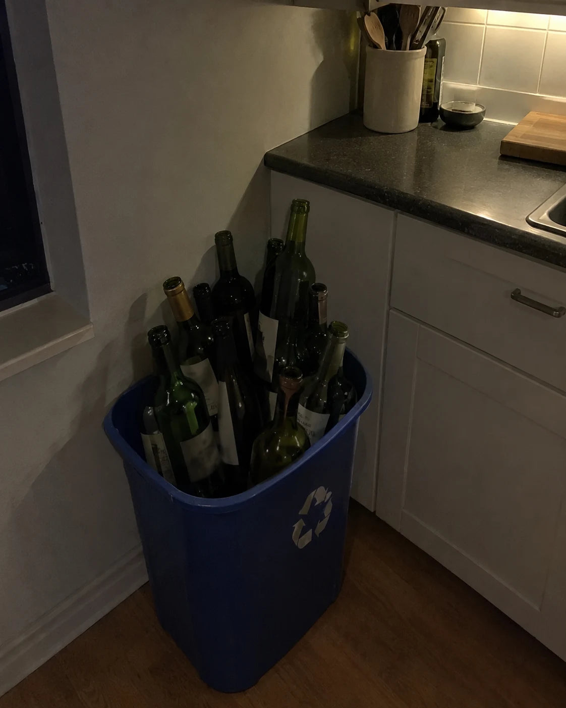 A recycling bin full of empty wine bottles in a dim kitchen at night, showing a repeated nightly pattern