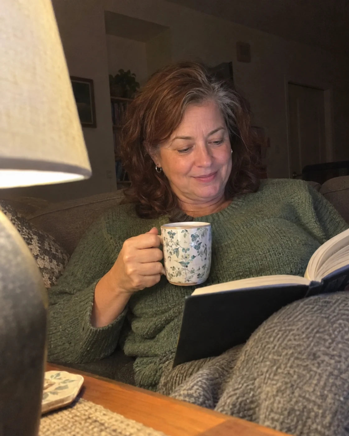 A woman relaxed on her couch in the evening with tea and a book, calm expression