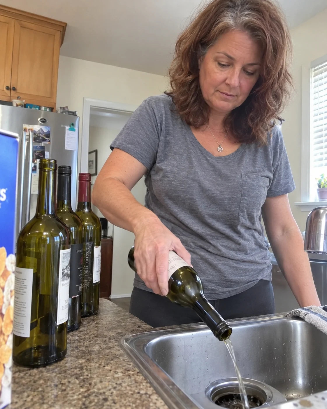 A woman pouring a wine bottle down the kitchen sink, phone propped on the counter filming her