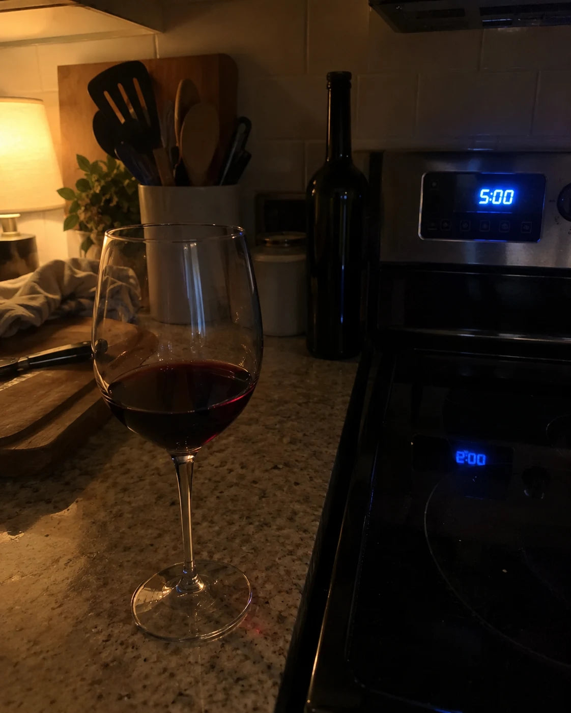 A wine glass left on a kitchen counter at night beside a stove clock reading 5:00