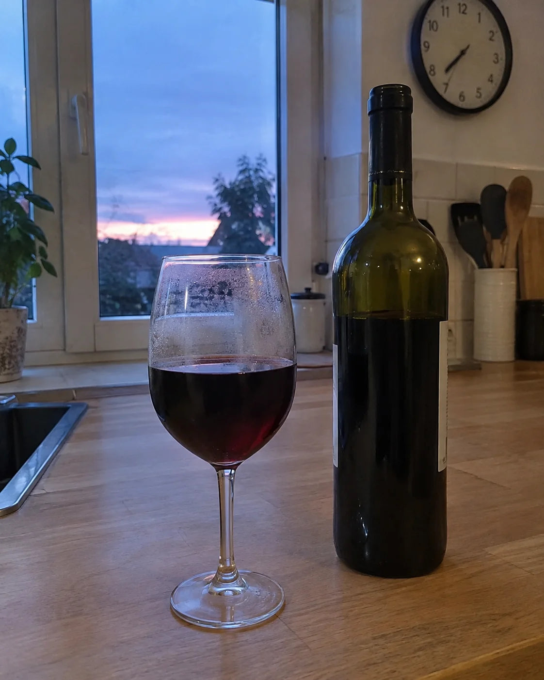 A full glass of red wine sitting untouched next to a bottle on a kitchen counter at dusk, a clock reading just past 7