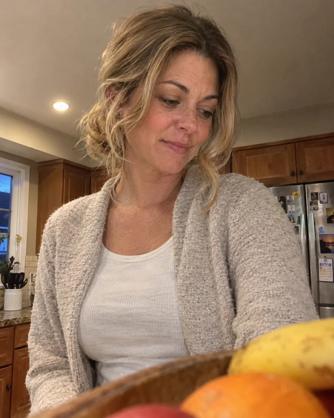 A woman standing at her kitchen counter in the evening, looking down thoughtfully, phone propped against a fruit bowl