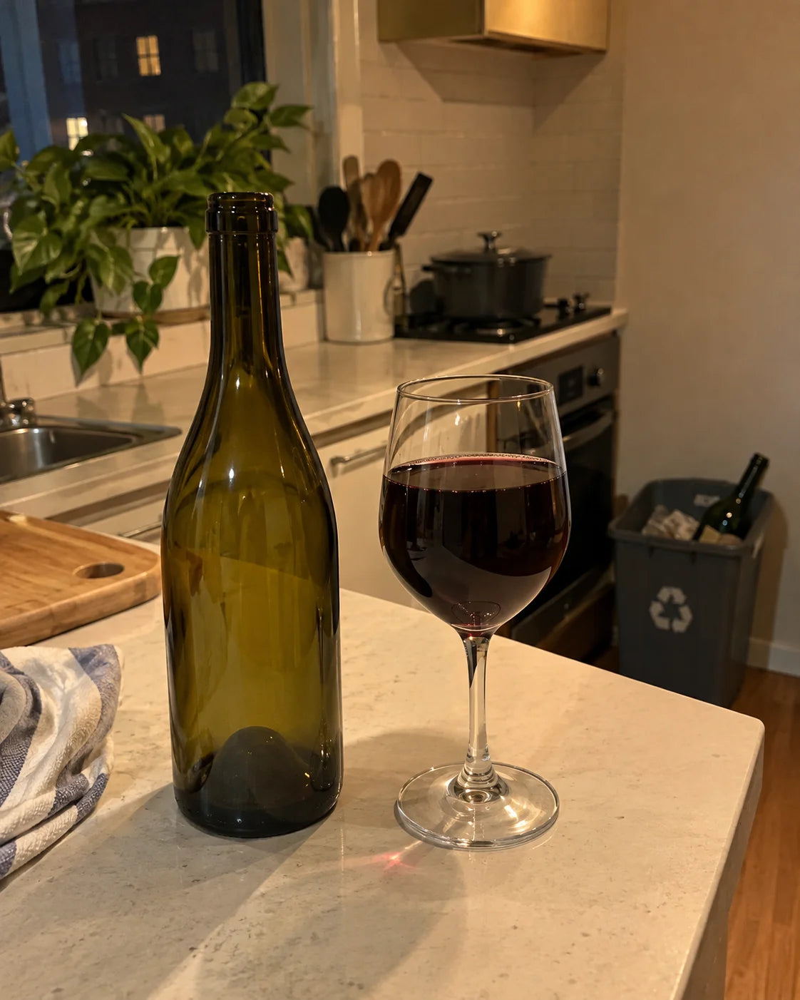 An empty wine bottle next to a nearly full wine glass on a kitchen counter at night