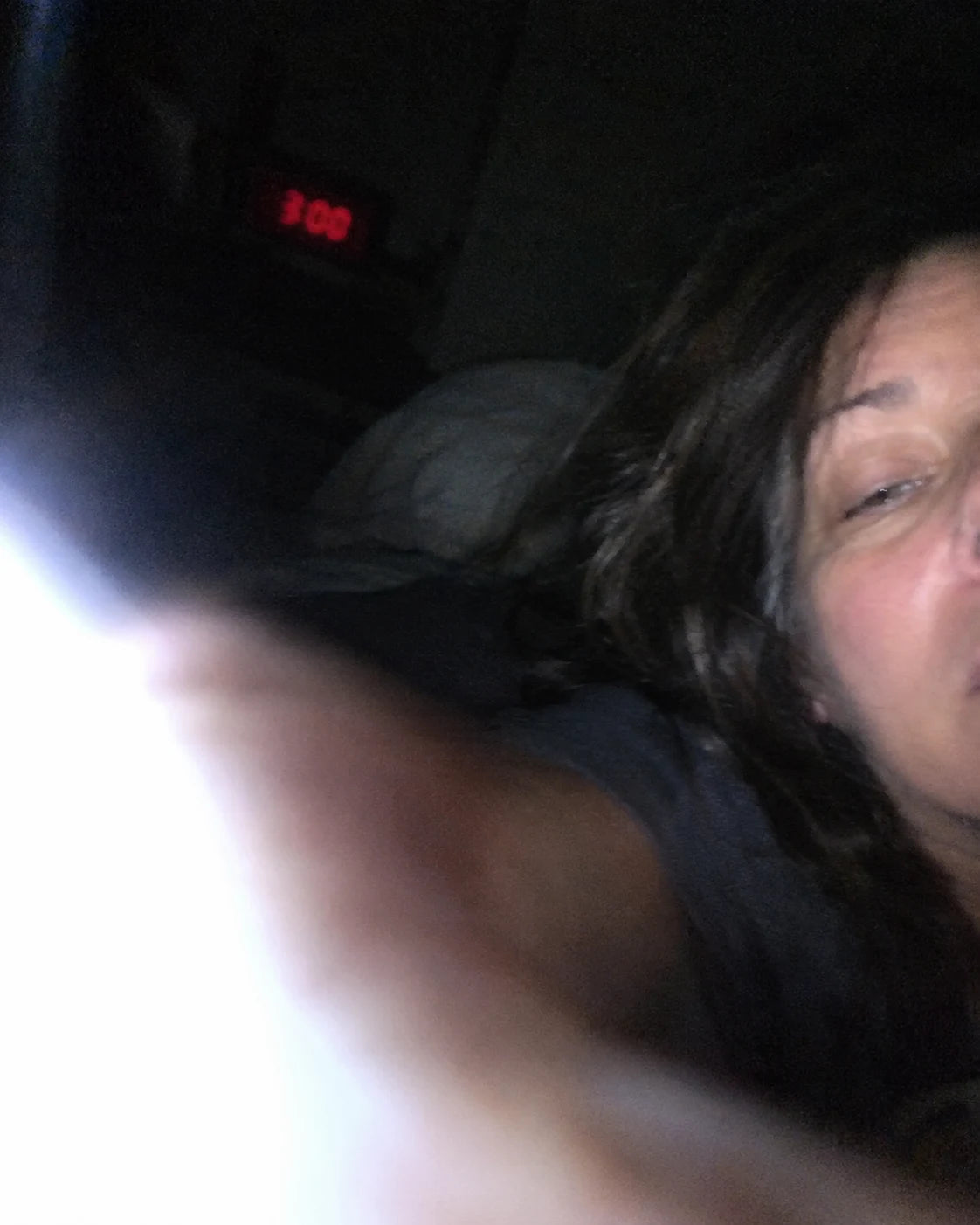 A woman lying awake in bed at night, checking the time on her phone, a digital clock on the nightstand reading 3:00