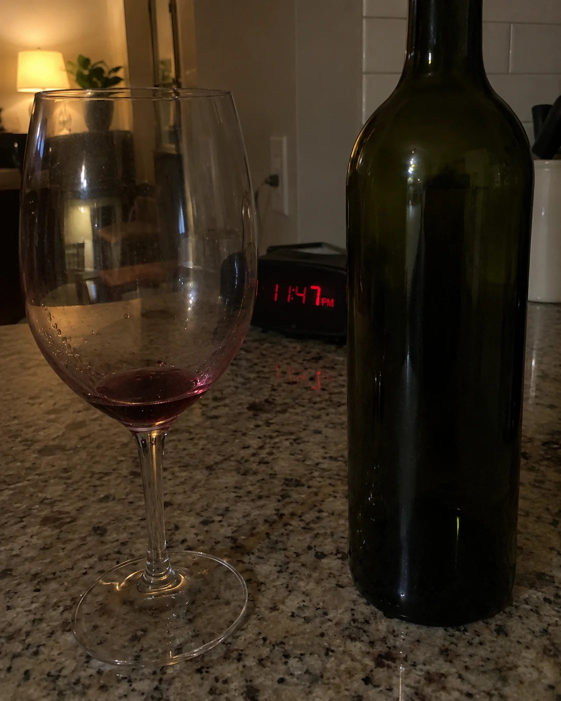 An empty wine glass on a kitchen counter at night next to a clock reading 11:47pm