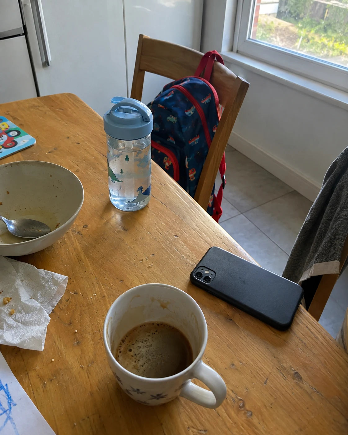 A cluttered kitchen table in the morning, a half-finished cup of coffee, a phone, a child's backpack on a chair in the background