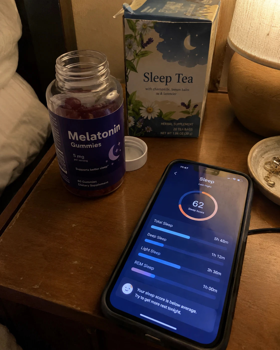 A nightstand at night with a bottle of melatonin gummies, a box of sleep tea, and a phone showing a sleep tracking app with a low sleep score