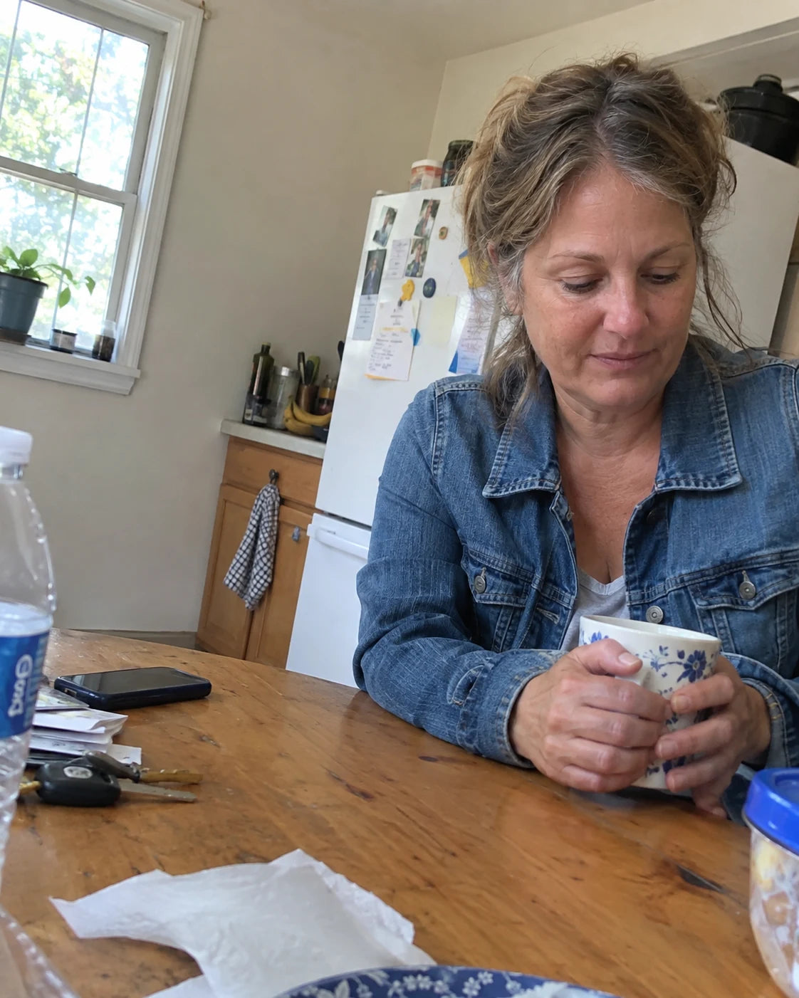 Candid off-center photo of her sitting at her kitchen table holding a coffee mug, looking down