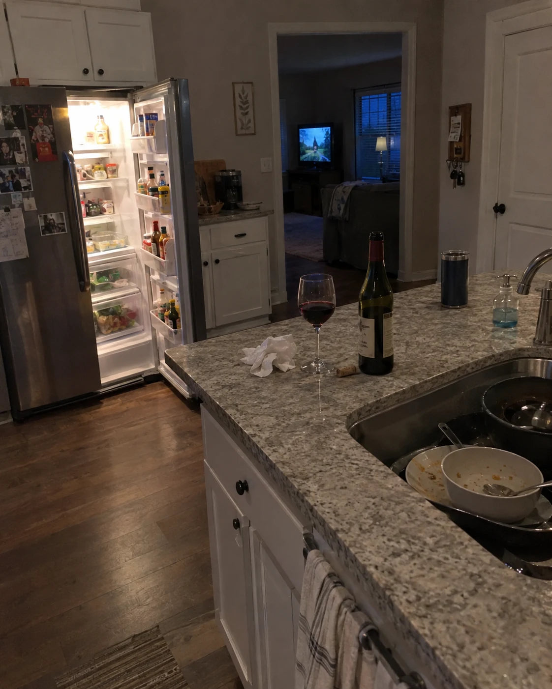 An open refrigerator glowing in a dim kitchen at dusk, a half-poured glass of wine and open bottle left on the counter next to dirty dishes