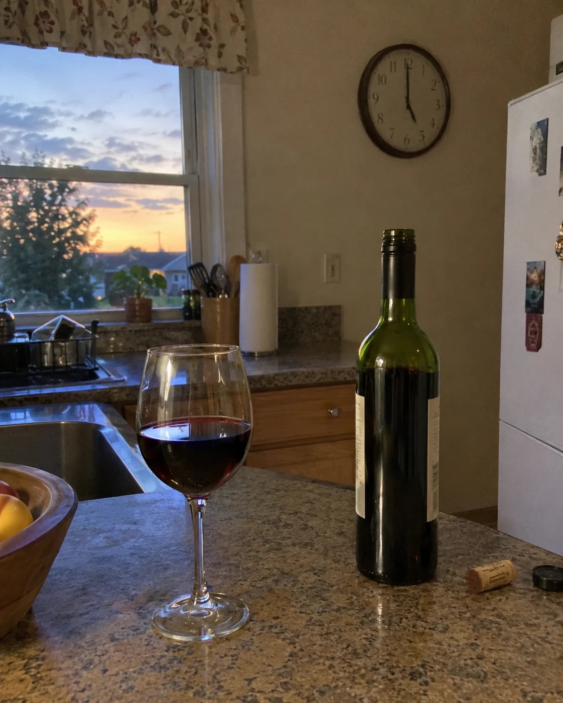 Kitchen counter at dusk with a poured glass of wine and a wall clock nearby, nobody in frame