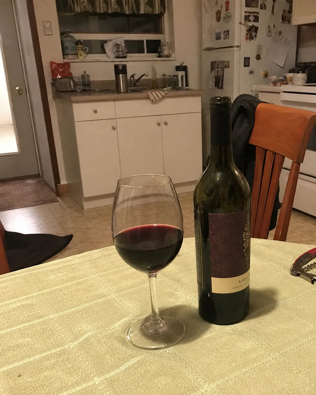 Glass of red wine next to an open bottle on a kitchen table in the evening