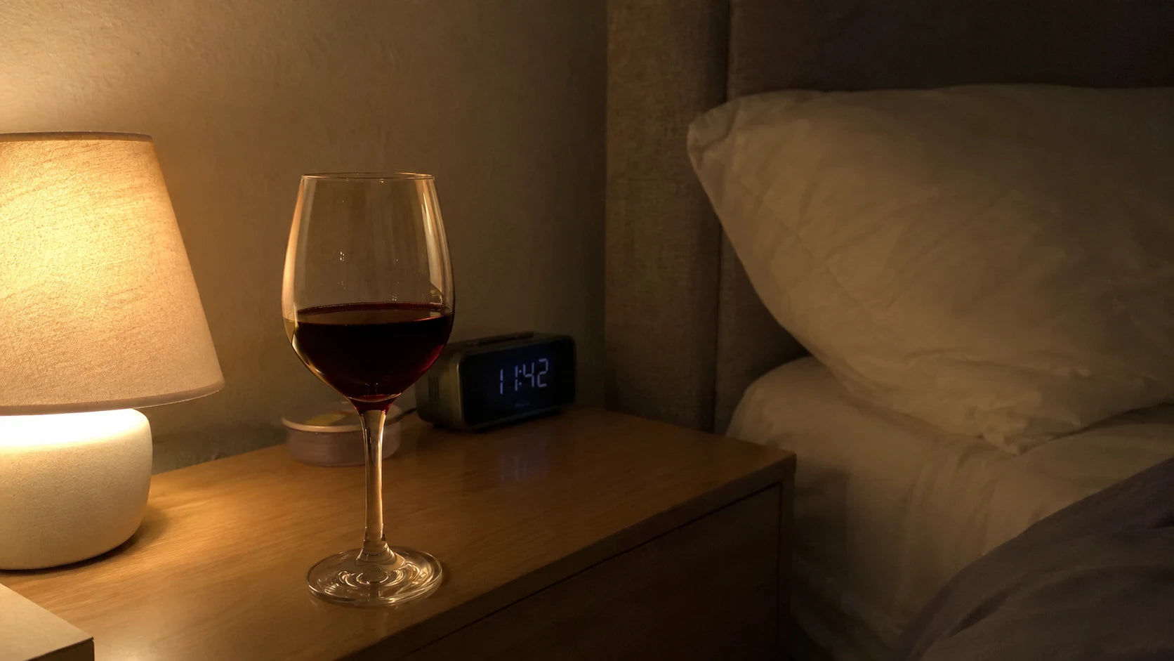 A wine glass on a nightstand beside a dim bedside lamp at night