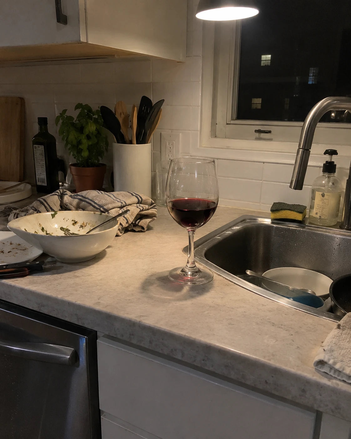 A wine glass left on a kitchen counter at night next to a cluttered sink