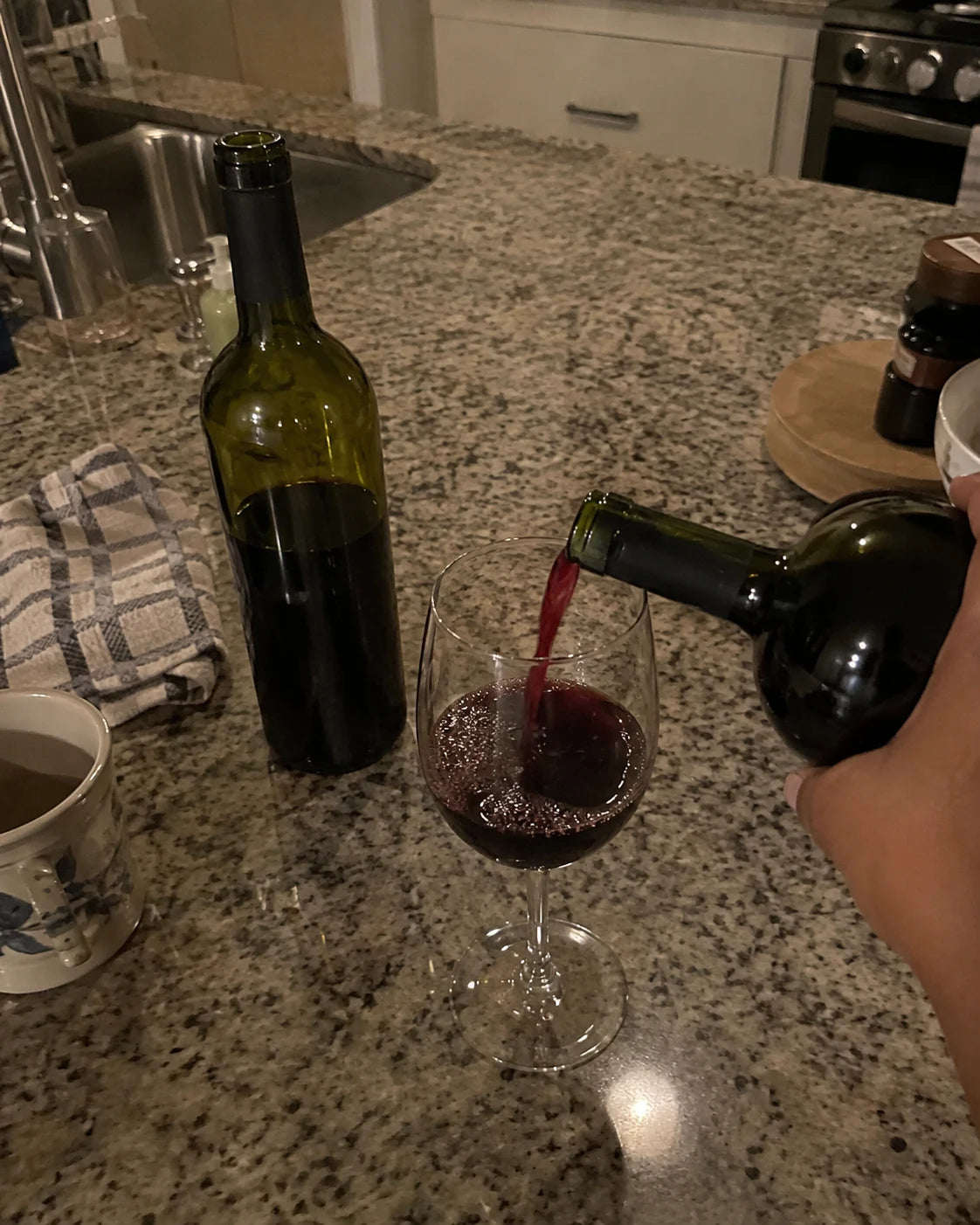 Hand pouring a second glass of wine at a kitchen counter at night