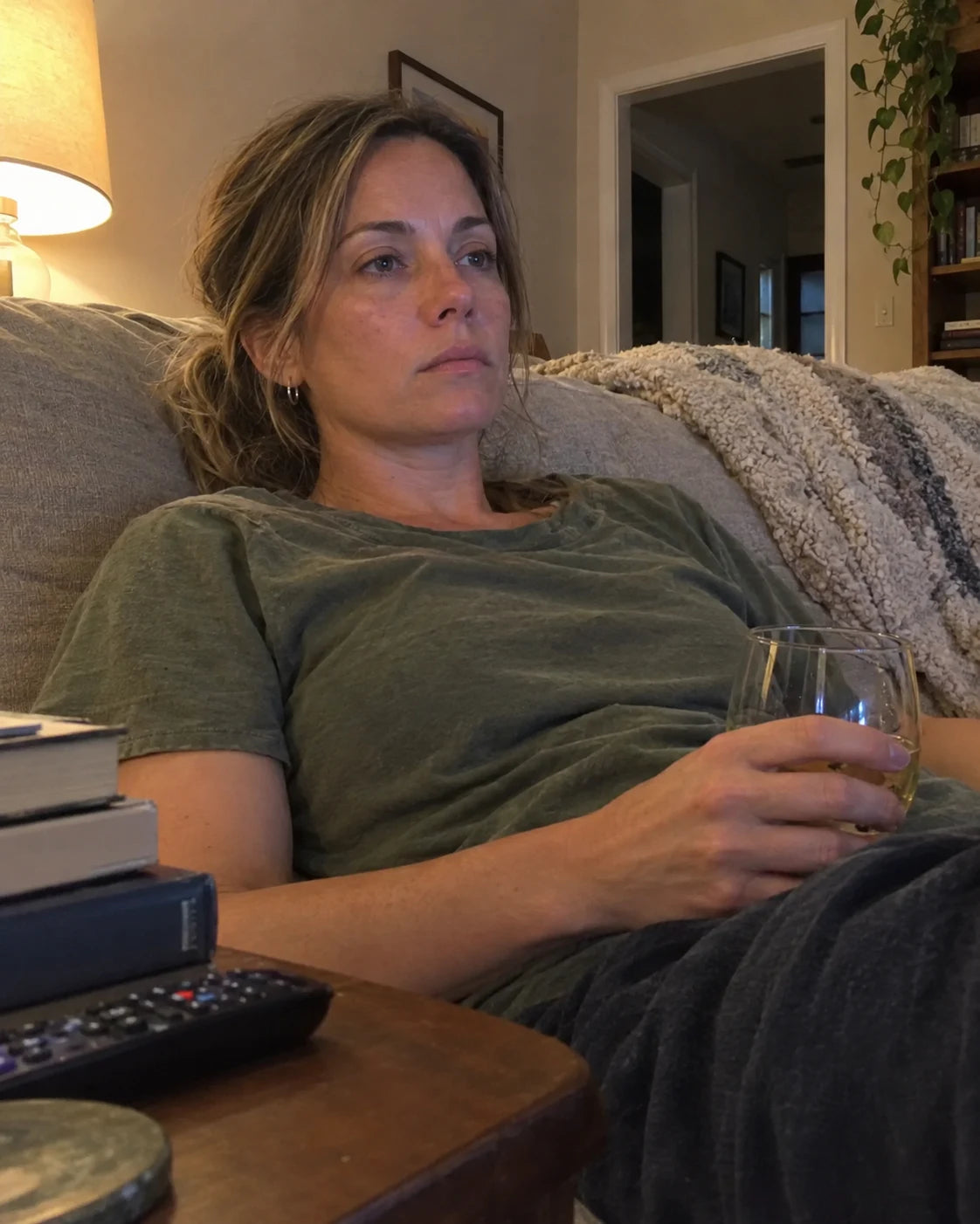 Woman reaching for a glass on the side table without looking, candid living room shot at dusk, phone propped on a stack of books