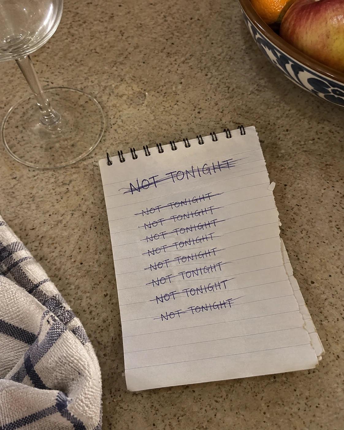 Notepad on a kitchen counter with 'not tonight' written and crossed out repeatedly, empty wine glass nearby